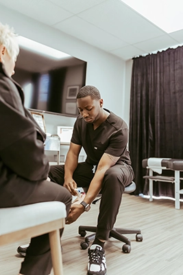 Person seated on a rolling stool applying a device to another person's lower leg in a clinical room.