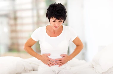 Person in a white shirt holding both hands over the abdominal area while seated on a bed.