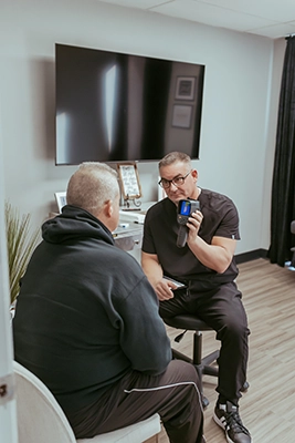 Person seated on a stool holding a handheld device while speaking with another seated person in a clinic room.