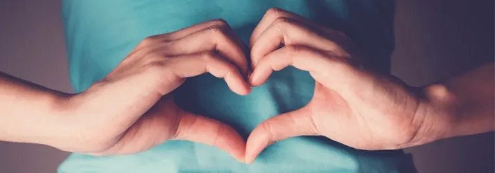 Close-up of two hands forming a heart shape against a blue shirt background.