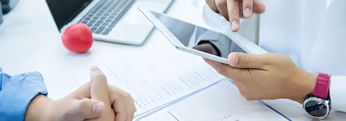 Copilot said: Close-up of a person holding a tablet and pointing at the screen during a consultation, with documents on a desk.