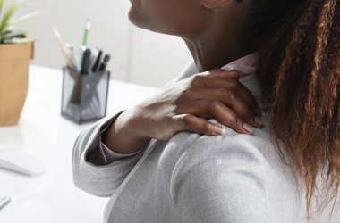 Close-up of a person touching the shoulder and upper back area while seated near a desk.