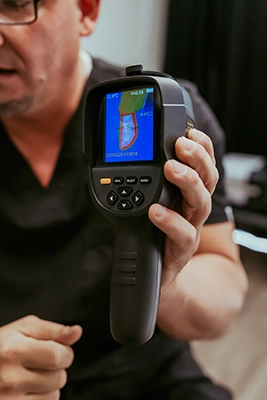 Close-up of a handheld thermal imaging device displaying a color heat map, held in front of a seated person.